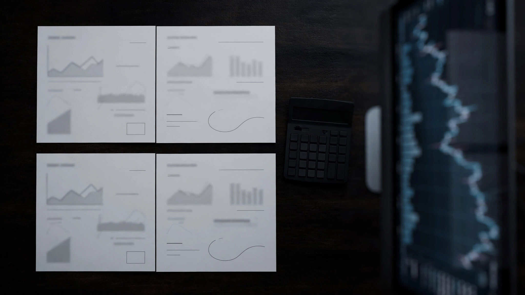 Top-down view of printed spread comparison sheets and a calculator beside a blurred EUR/USD chart